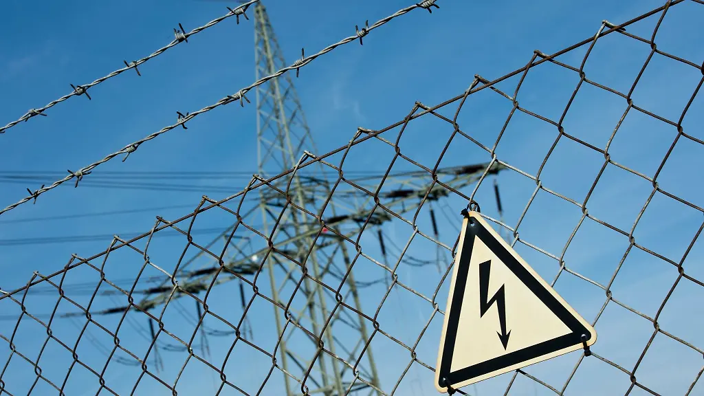 Ein-Gefahrenschild-fuer-Elektrizitaet-haengt-vor-einer-Stromtrasse-an-einem-Zaun