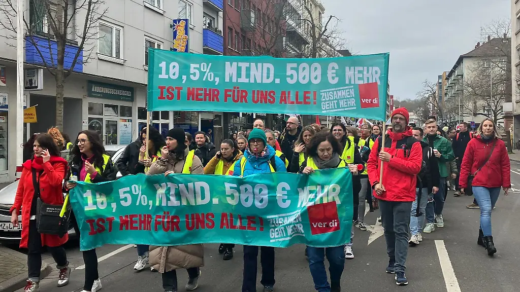Teilnehmer-und-Teilnehmerinnen-eines-Warnstreiks-in-Mainz-halten-Banner-mit-der-Aufschrift-10-5-mind-500-Euro-mehr-ist-mehr-fuer-uns-alle-in-die-Hoehe