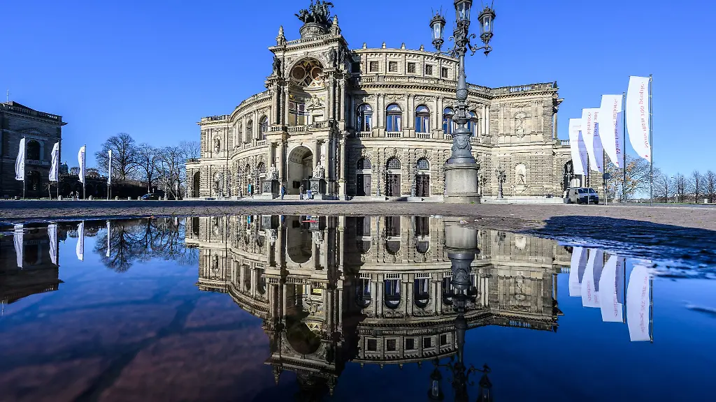 Die-Semperoper-spiegelt-sich-bei-Sonnenschein-in-einer-Pfuetze-auf-dem-Theaterplatz