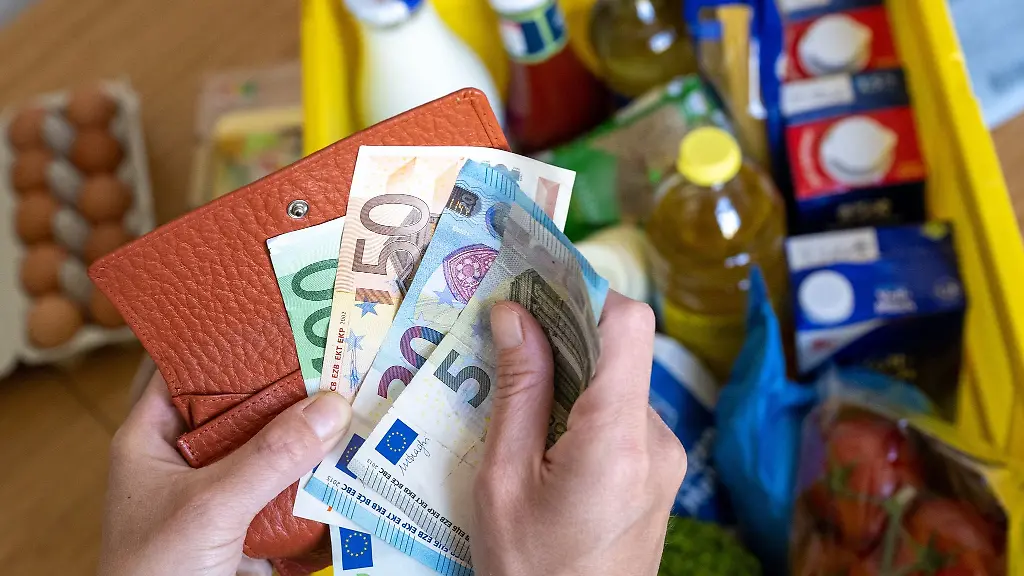 Eine-Einkaufskiste-mit-Lebensmitteln-steht-auf-einem-Kuechentisch-waehrend-eine-Frau-Euro-Banknoten-in-den-Haenden-haelt