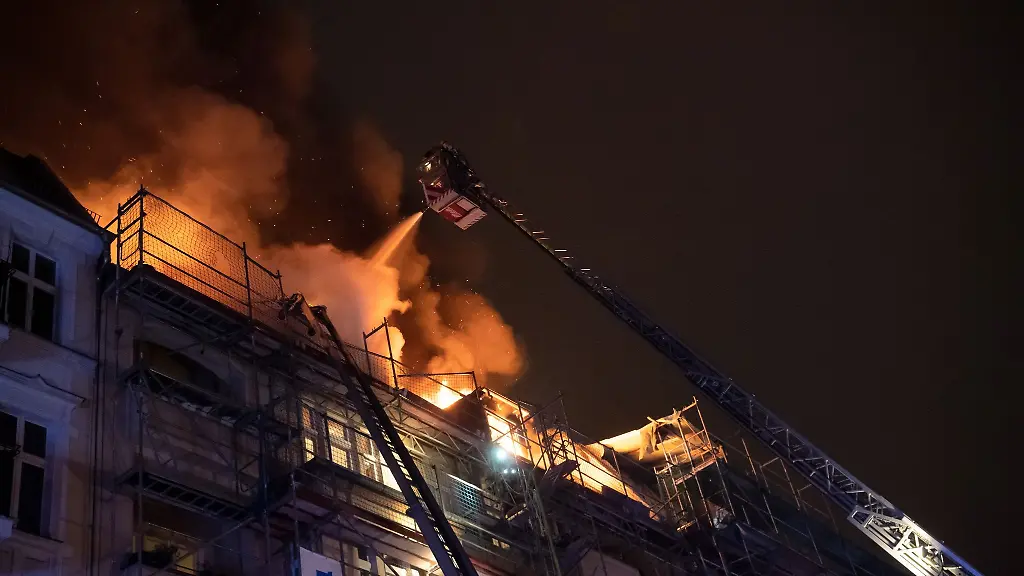 Ein-Feuerwehrmann-loescht-von-einer-Drehleiter-aus-die-Flammen-auf-dem-Dach-eines-Wohnhauses