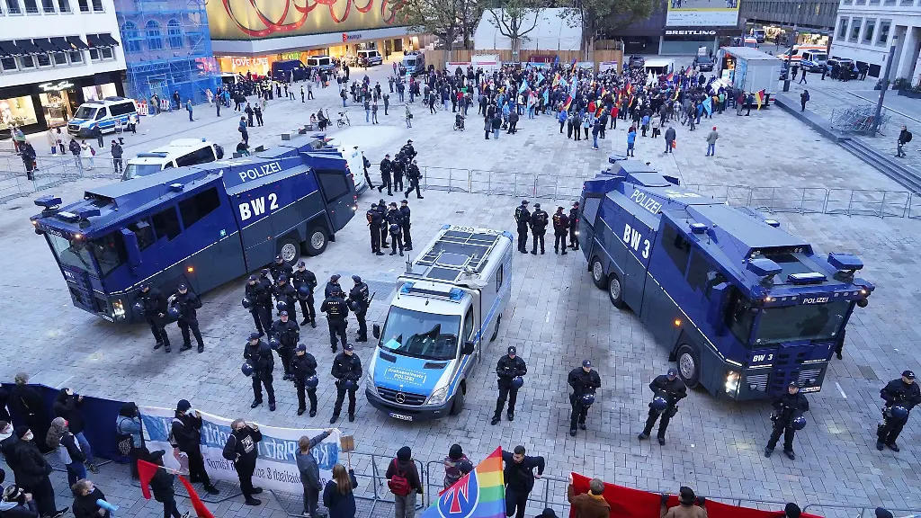 Polizisten-stehen-mit-Wasserwerfern-am-Marktplatz-vor-Demonstranten-vorne-unten-die-gegen-eine-AfD-Kundgebung-im-Hintergrund-protestieren