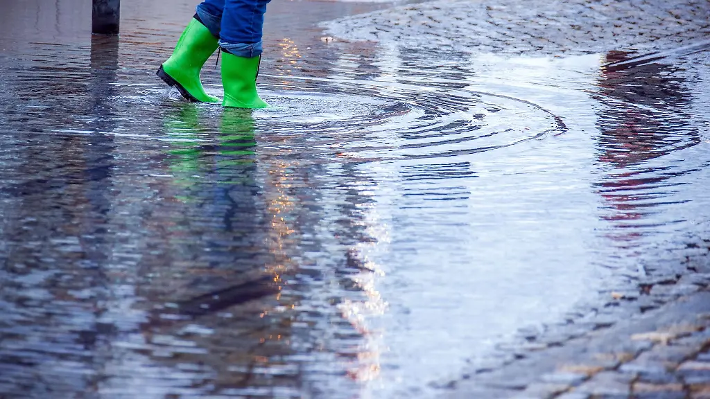 Mit-Gummistiefeln-geht-eine-Frau-ueber-eine-leicht-ueberflutete-Strasse