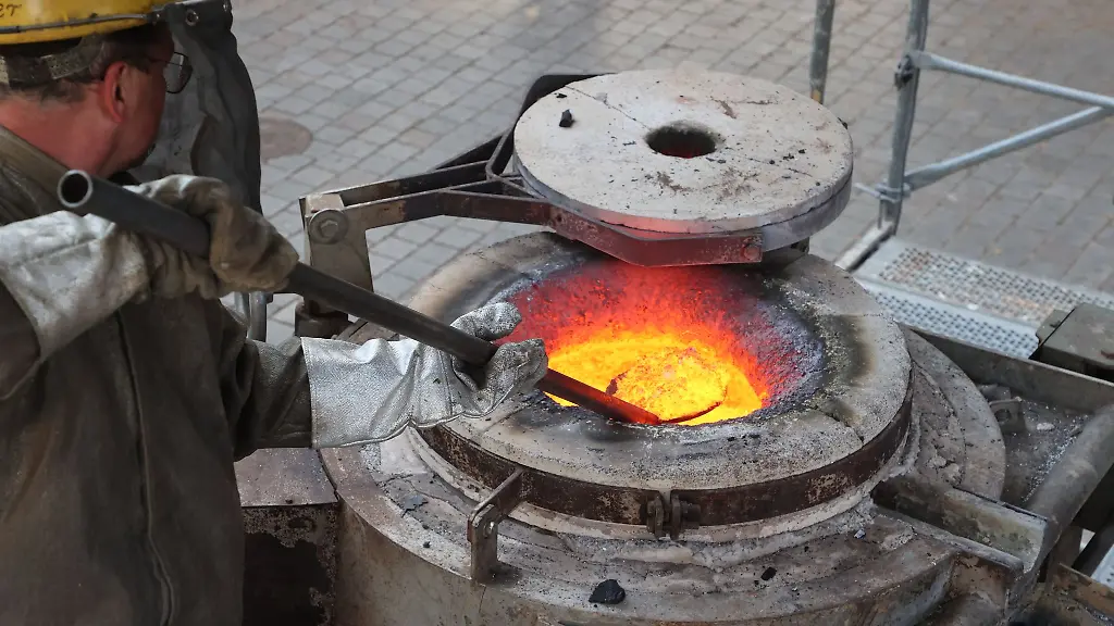 Mitarbeiter-einer-Glockenbaufirma-arbeiten-an-einem-Ofen-in-welchem-Metall-erhitzt-wird-und-dann-ueber-einen-Steg-in-fluessiger-Form-in-die-Huelle-der-neuen-Glocke-gegossen-wird