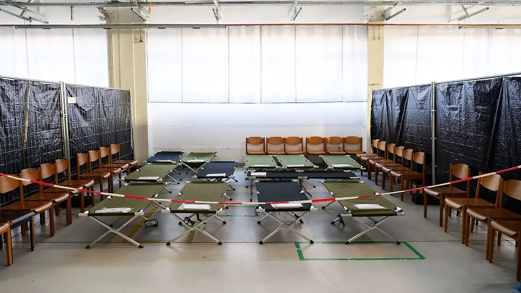 Feldbetten-und-Holzstuehle-stehen-in-einer-Halle