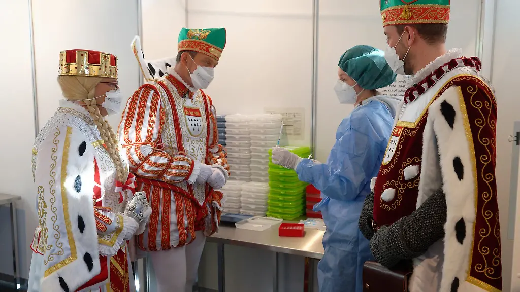 Eine-Mitarbeiterin-erklaert-dem-Koelner-Dreigestirn-an-einer-mit-Wasser-gefuellten-Flasche-die-Dosierung-von-Impfstoff