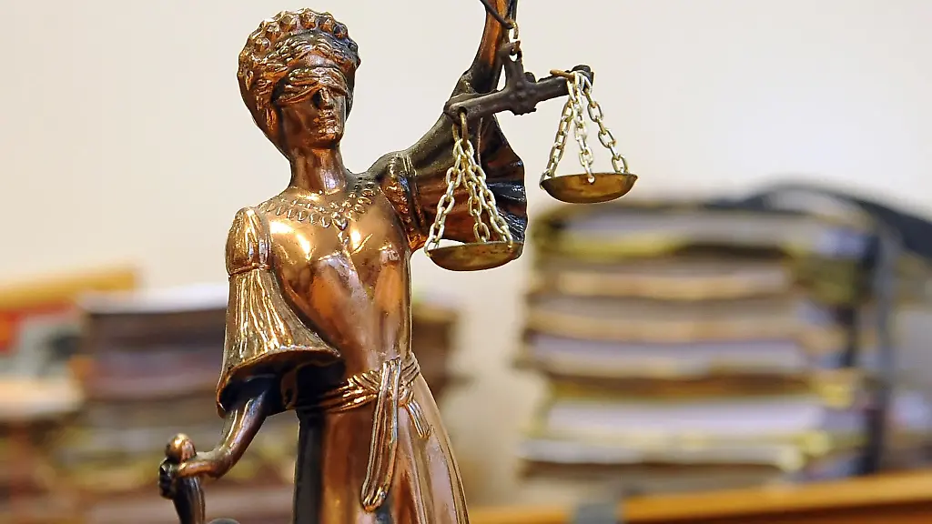 Eine-goldfarbene-Justitia-Figur-steht-vor-Aktenbergen-die-sich-auf-einem-Tisch-stapeln