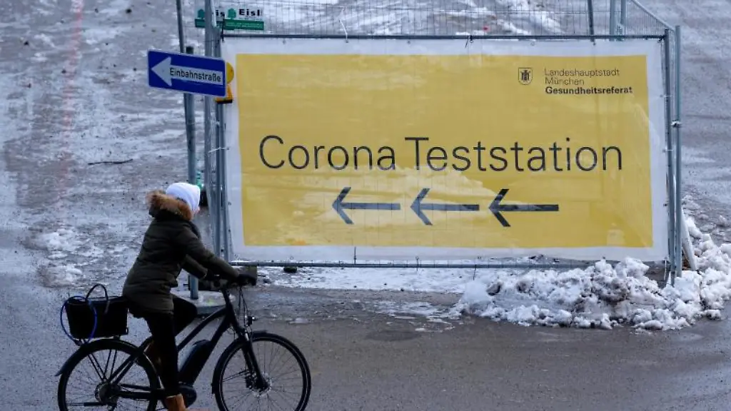 Eine-Frau-faehrt-mit-ihrem-Fahrrad-an-einem-Plakat-mit-der-Aufschrift-Corona-Teststation-entlang