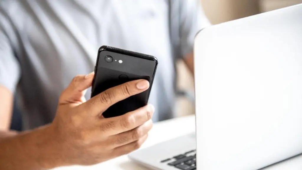 Ein-Mann-sitzt-vor-einem-Laptop-und-haelt-ein-Handy-in-der-Hand