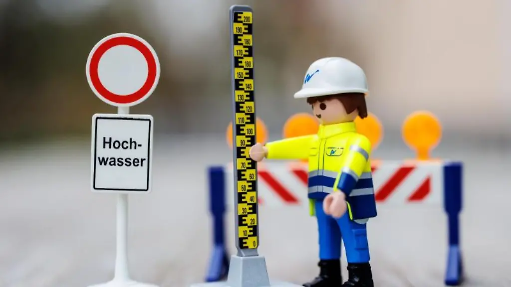 Eine-Playmobil-Figur-mit-einer-Pegellatte-einer-Absperrschranke-und-einem-Schild-mit-der-Aufschrift-Hochwasser-steht-auf-einem-Tisch