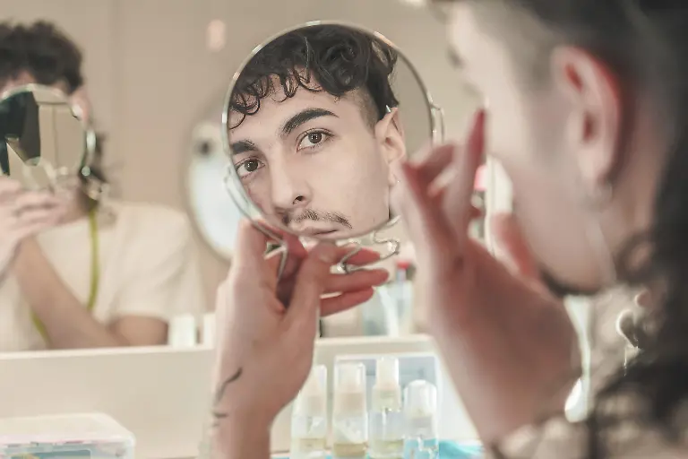 Junger-Mann-der-sein-Gesicht-im-Spiegel-betrachtet-waehrend-er-im-Badezimmer-Cremes-fuer-seine-taegliche-Hautpflege-auftraegt-mit-einer-anderen-unscharfen-Person-im-Hintergrund-Modellfreigabe-vorhanden