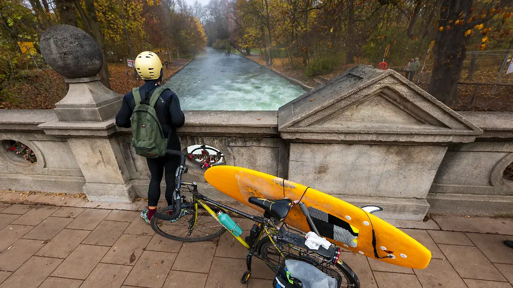 Ein-Mann-mit-einem-Surfboard-an-seinem-Fahrrad-schaut-von-einer-Bruecke-auf-die-Eisbachwelle-im-Englischen-Garten-die-seit-Tagen-verschwunden-ist-und-jetzt-repariert-werden-soll-Die-Welle-ist-zu-jeder-Jahres-und-Tageszeit-ein-wahrer-Hotspot-fuer-Touristen-und-Surfer-aus-der-ganzen-Welt