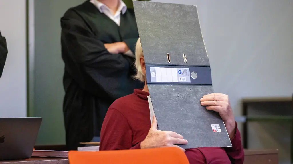 Ein-77-jaehriger-Angeklagter-sitzt-zum-Prozess-Auftakt-in-einem-Sitzungssaal-im-Landgericht-Muenchen-I-und-haelt-sich-einen-Aktenordner-vor-das-Gesicht-Der-angeklagte-77-Jaehrige-soll-die-Nichte-seiner-Frau-aus-einem-Waisenhaus-in-Brasilien-geholt-und-sie-dann-ueber-Jahre-schwer-sexuell-missbraucht-haben