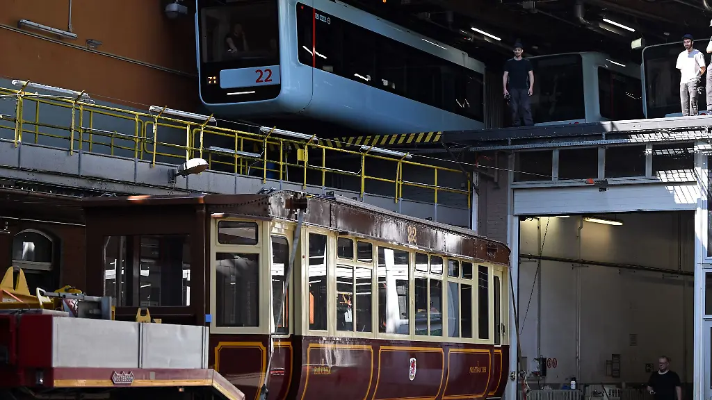 Die-Schwebebahn-gestern-und-heute-Ein-Wagen-der-ersten-Baureihe-1900-steht-auf-einem-Tieflader-unter-einem-Wagen-der-aktuellen-Baureihe-in-der-Werkstatt-der-Schwebebahn