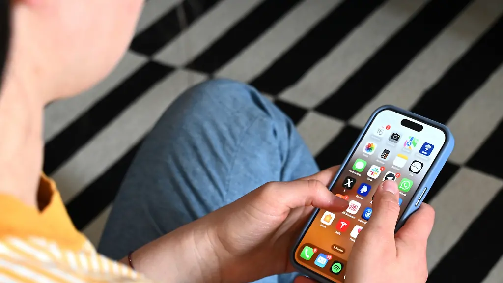 Eine-Jugendliche-sitzt-auf-dem-Boden-und-nutzt-ein-Smartphone-auf-dem-Display-sind-verschiedene-Apps-zu-sehen-In-Australien-wird-das-Social-Media-Verbot-fuer-Kinder-unter-16-Jahren-um-zwei-Plattformen-erweitert-Ab-dem-10-Dezember-muessen-sich-auch-das-Internetforum-Reddit-und-die-australische-Livestreaming-Plattform-Kick-com-an-die-neuen-gesetzlichen-Vorgaben-halten