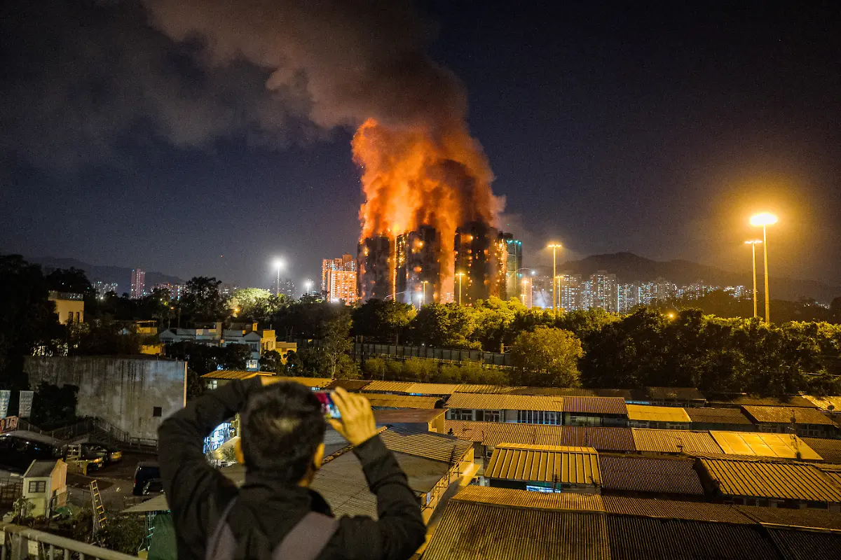 An-onlooker-takes-photos-as-a-major-fire-engulfs-several-apartment-blocks-at-the-Wang-Fuk-Court-residential-estate-background-in-Hong-Kong-s-Tai-Po-district-on-November-26-2025-At-least-four-people-were-killed-when-a-fire-engulfed-several-high-rise-blocks-in-a-Hong-Kong-residential-estate-on-November-26-the-government-said-with-media-reporting-that-some-residents-were-trapped-inside
