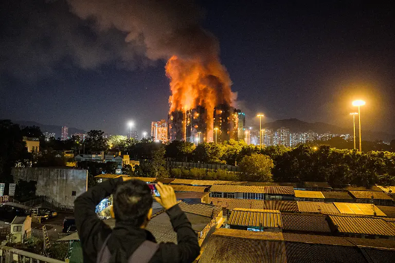An-onlooker-takes-photos-as-a-major-fire-engulfs-several-apartment-blocks-at-the-Wang-Fuk-Court-residential-estate-background-in-Hong-Kong-s-Tai-Po-district-on-November-26-2025-At-least-four-people-were-killed-when-a-fire-engulfed-several-high-rise-blocks-in-a-Hong-Kong-residential-estate-on-November-26-the-government-said-with-media-reporting-that-some-residents-were-trapped-inside