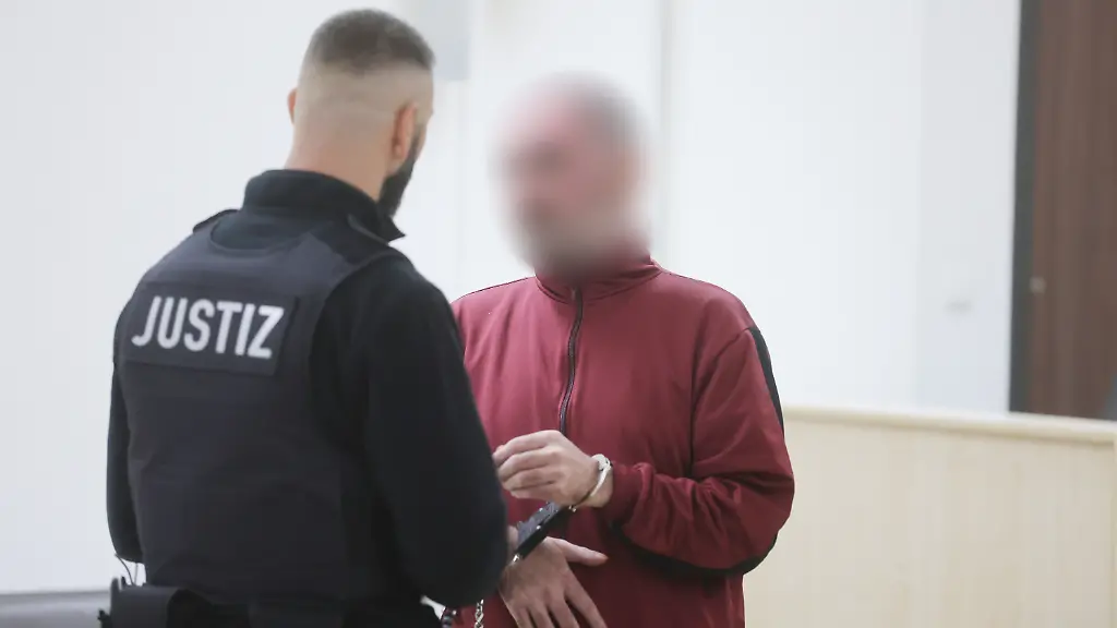 Der-Angeklagte-r-wird-von-einem-Justizbeamten-in-den-Gerichtssaal-gefuehrt-Dem-46-Jahre-alten-Mann-wird-vorgeworfen-dass-er-seine-Frau-in-einer-Strassenbahn-mit-Benzin-uebergossen-und-dann-angezuendet-haben-soll-Die-Frau-wurde-bei-der-Tat-am-16-Maerz-2025-lebensgefaehrlich-verletzt