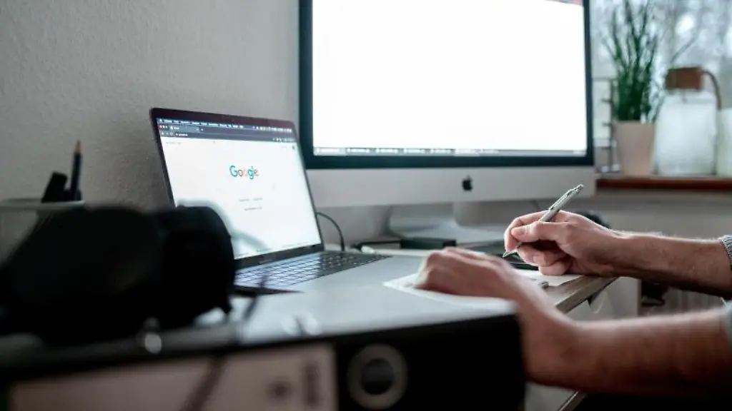 Ein-Mann-sitzt-mit-einem-Laptop-und-einem-Bildschirm-an-einem-Tisch-vor-einem-Fenster-im-Homeoffice