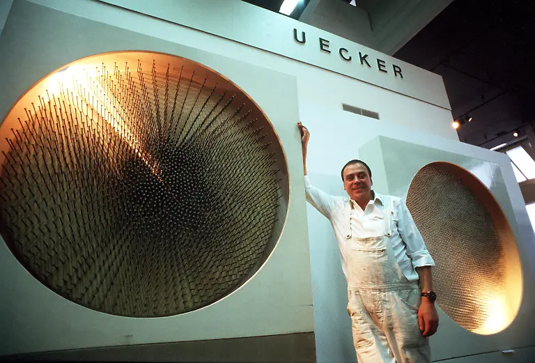 Der-deutsche-Bildhauer-und-Objektkuenstler-Guenther-Uecker-steht-am-12-12-1987-neben-seinem-Werk-Lichtscheibe-aus-dem-Jahr-1970-das-im-Rahmen-einer-Ausstellung-in-Ludwigshafen-gezeigt-wird-Seit-1958-ist-der-in-Wendorf-geborene-Uecker-als-freischaffender-Kuenstler-taetig-Er-gehoert-als-Mitglied-der-Gruppe-Zero-zu-den-profiliertesten-und-vielseitigsten-Vertretern-der-deutschen-Nachkriegskunst-Sein-Markenzeichen-ist-der-Nagel-mit-dem-er-immer-neue-Wege-wagte-Von-Scheiben-organischen-Formationen-bis-hin-zu-uebernagelten-Moebelstuecken-reichen-seine-in-die-Kunstgeschichte-eingegangenen-Nagelbilder-Internationale-Anerkennung-fand-der-in-Duesseldorf-lebende-und-lehrende-Professor-fuer-Freie-Kunst-durch-Projekte-in-Nordamerika