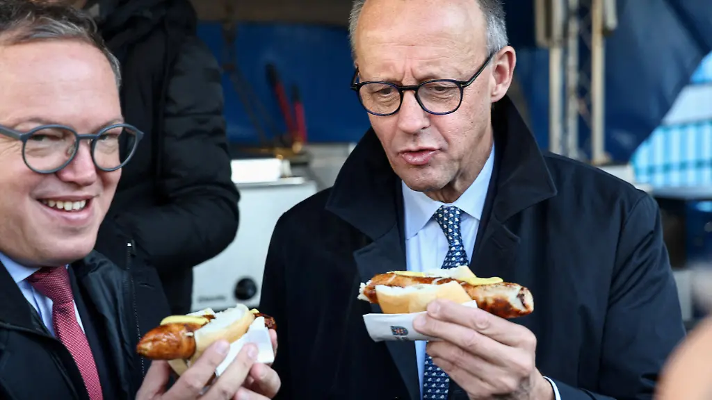 German-Chancellor-Friedrich-Merz-and-Thuringia-s-state-premier-Mario-Voigt-each-hold-a-sausage-in-a-bun-during-Merz-s-visit-to-a-school-in-Jena-Lobeda-in-Thuringia-Germany-December-2-2025