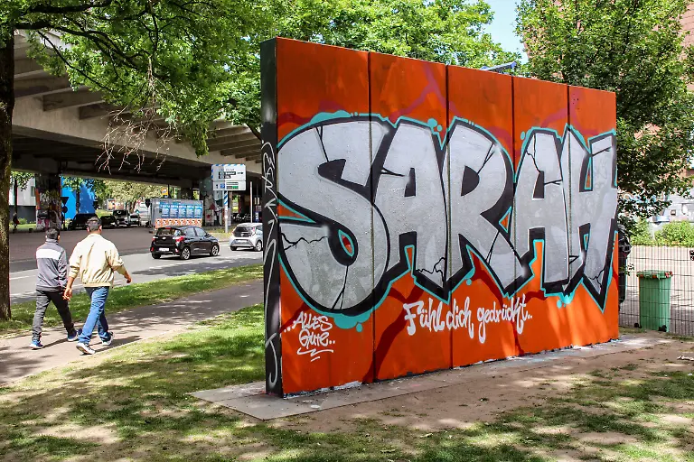 Ein-Graffiti-mit-dem-Namen-Sarah-in-einer-Gruenanlage-in-Bielefeld-zum-Gedenken-an-eine-junge-Frau-die-bei-einer-Messerattacke-vor-der-Cutie-Bar-am-Sonntag-dem-18-05-2025-zusammen-mit-drei-anderen-jungen-Menschen-mutmasslich-von-einem-35-jaehrigen-Syrer-schwer-verletzt-wurde-Foto-vom-23-05-2025-Knapp-eine-Woche-nach-dem-Messerangriff-in-Bielefeld-hat-eine-Gruppierung-aus-dem-rechten-Spektrum-fuer-Samstag-einen-Aufmarsch-mit-Mahnwache-in-der-Stadt-angekuendigt-Dazu-wuerden-200-Teilnehmer-erwartet-teilte-die-Polizei-Bielefeld-am-Freitag-mit-Die-angemeldete-Versammlung-soll-vor-dem-Tatort-der-Tanzbar-Cuties-stattfinden-Das-Motto-der-rechtsgerichteten-Kundgebung-lautet-Anteilnahme-fuer-die-Opfer-vom-18-05-2025-und-Remigration-Ein-Bielefelder-Buendnis-rief-zu-einer-Gegendemonstration-unter-dem-Motto-Gegen-Hass-und-Hetze-in-Bielefeld-auf-Dazu-sind-laut-Polizei-500-Teilnehmerinnen-und-Teilnehmer-angemeldet-Man-sei-mit-verstaerkten-Kraeften-im-Einsatz-und-gehe-von-einem-friedlichen-Verlauf-der-Versammlungen-aus-erklaerte-das-Polizeipraesidium-Bielefeld-Siehe-epd-Meldung-vom-23-05