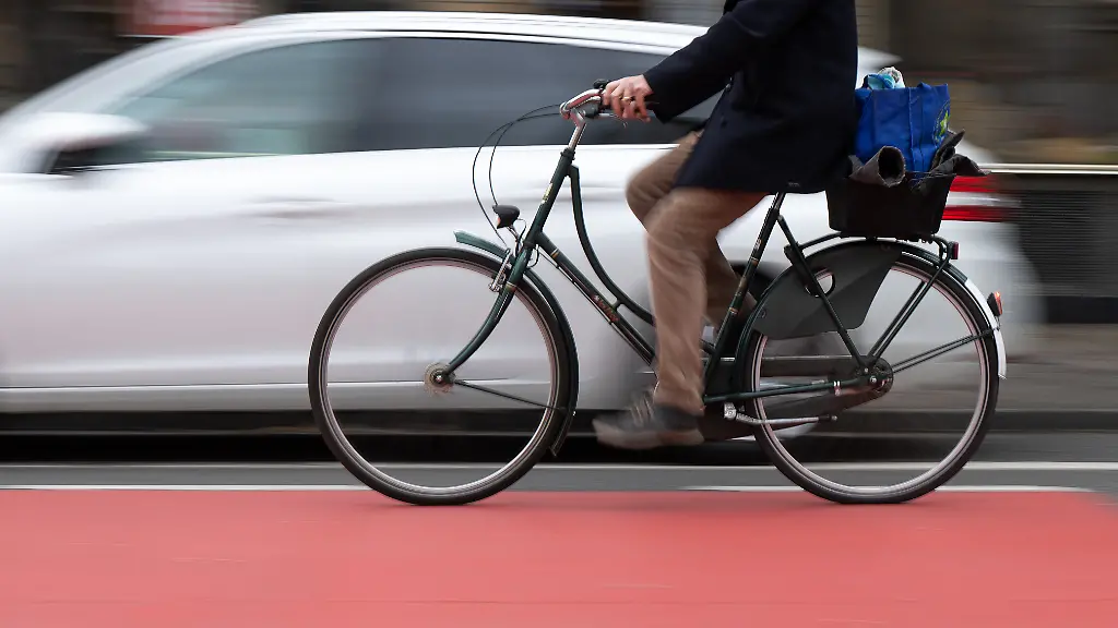 Ein-Fahrradfahrer-faehrt-auf-einer-Fahrradstrasse-an-einem-Schutzstreifen-fuer-geoeffnete-Tueren-an-Autos-vorbei-Wischeffekt-durch-Langzeitbelichtung-Der-Schutzstreifen-soll-verhindern-dass-Fahrradfahrer-durch-unachtsam-geoeffnete-Tueren-verletzt-werden