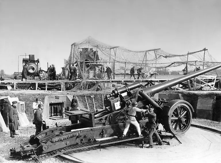 Eine-schwere-Feldhaubitze-18-waehrend-einer-Uebung-in-einer-offenen-Feuerstellung-am-Atlantikwall-Im-Hintergrund-wird-am-Ausbau-der-Stellung-gearbeitet-Aufnahmedatum-01-01-1942-31-12