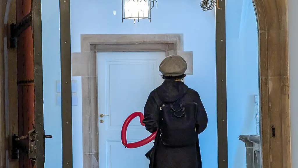 A-woman-holds-a-red-heart-shaped-balloon-and-stands-in-front-of-a-white-door-inside-a-historic-building-in-Munich-Bavaria-Germany-on-May-3-2025-The-image-centers-on-the-symbol-of-the-heart