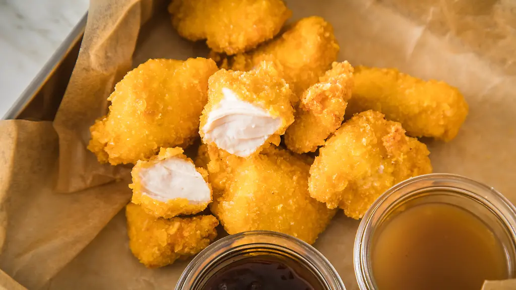 Chicken-Nuggets-liegen-neben-Sossen-auf-einem-Stueck-Backpapier-Sie-sind-verzehrfertig-ansprechend-verpackt-enthalten-oft-viel-Zucker-Salz-Fett-und-Zusatzstoffe-aber-wenig-wichtige-Naehrstoffe-Stark-verarbeitete-Lebensmittel-landen-sehr-haeufig-im-Einkaufswagen