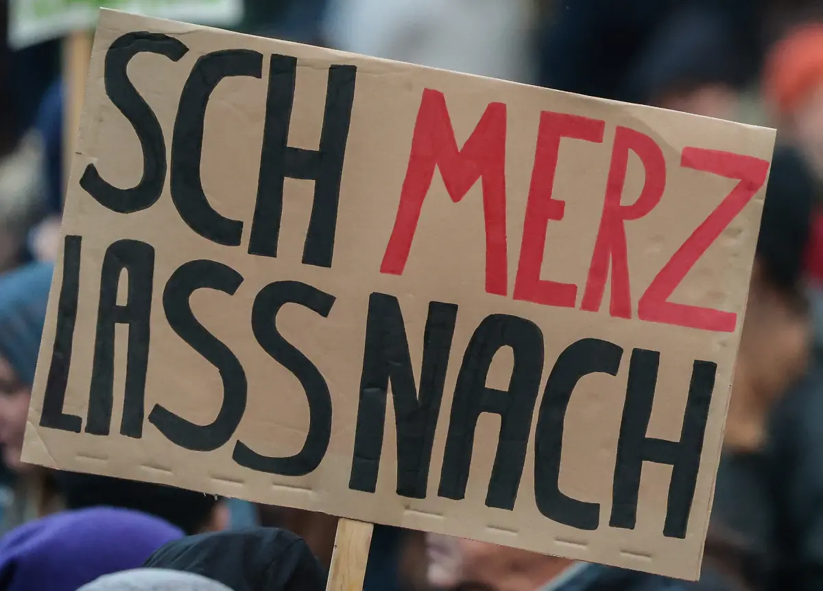 Ein-Schild-mit-der-Aufschrift-SchMerz-lass-nach-ist-bei-einer-Kundgebung-gegen-Aeusserungen-von-Bundeskanzler-Merz-auf-dem-Domshof-zu-sehen-Der-Protest-richtete-sich-gegen-Aussagen-von-Merz-zu-Stadtbild-und-Migration