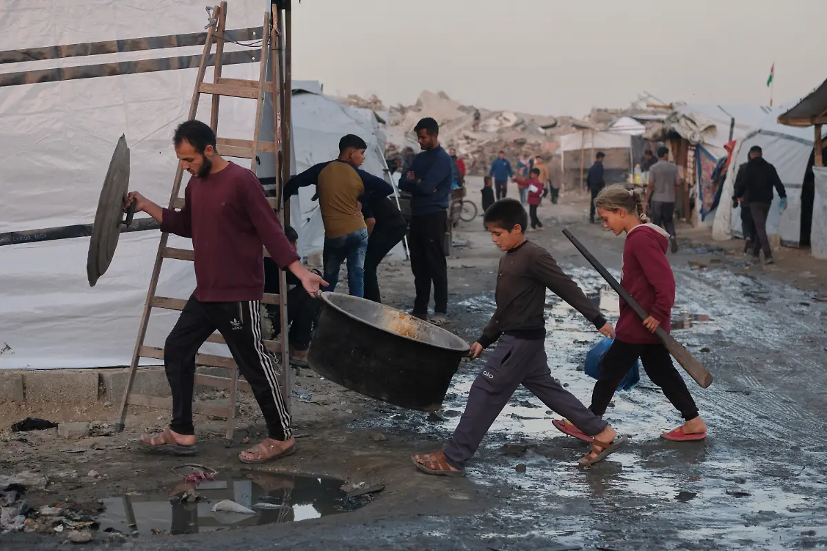 Vertriebene-Palaestinenser-gehen-durch-schlammige-Wege-waehrend-ein-Mann-und-Kinder-in-einem-Lager-in-Jabalia-im-noerdlichen-Gazastreifen-Kochausruestung-tragen