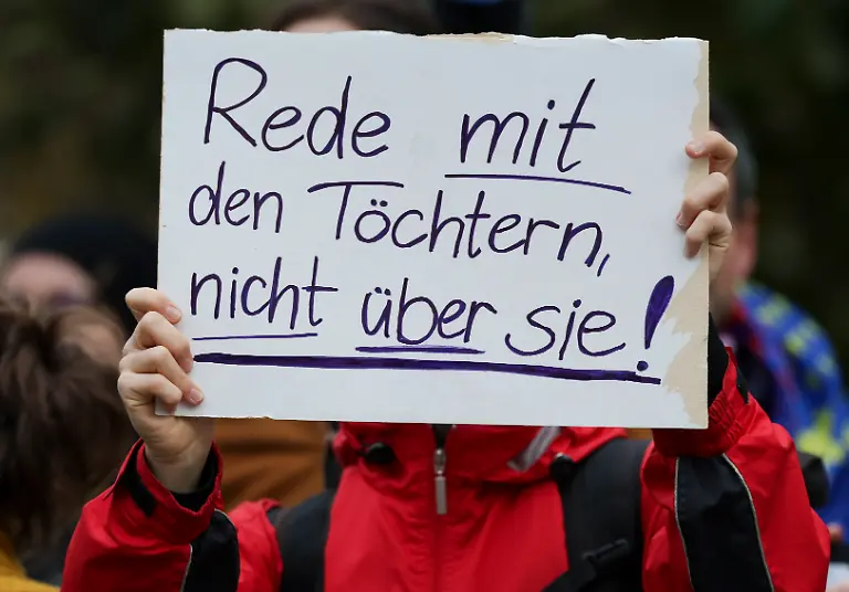 Eine-Demonstrantin-protestiert-mit-einem-Schild-mit-der-Aufschrit-Rede-mit-den-Toechtern-nicht-ueber-sie-gegen-des-Stadtbild-Zitat-waehrend-des-Antrittsbesuchs-gegen-Bundeskanzler-Merz-vor-der-saechsischen-Staatskanzlei