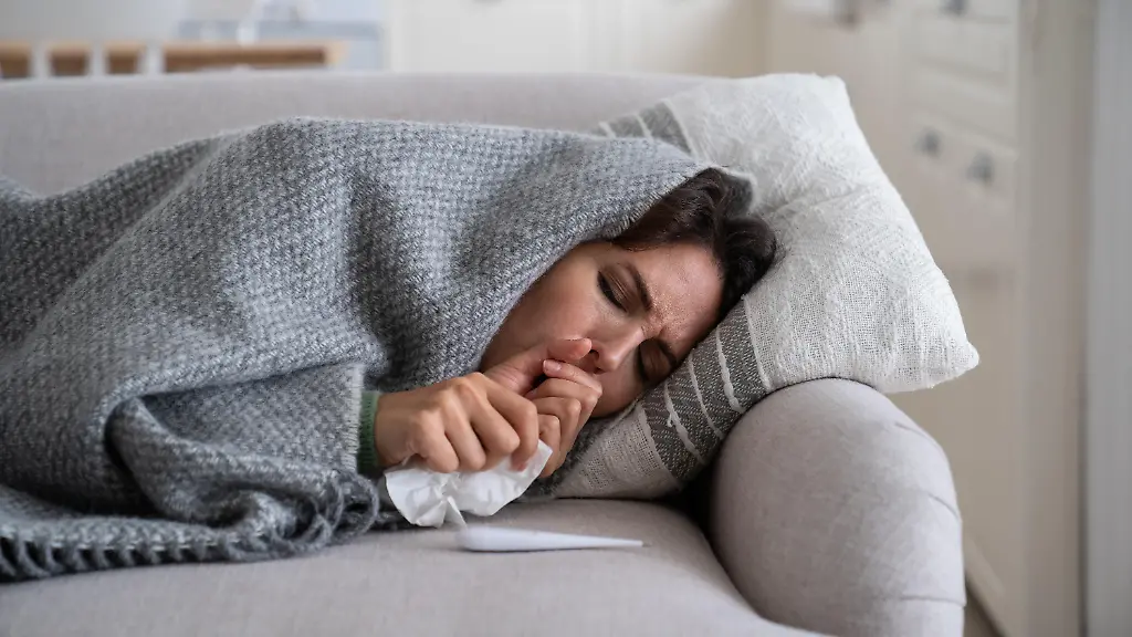 Young-woman-lying-under-blanket-on-sofa-coughing-and-sneezing-using-paper-tissue-to-blow-her-nose-Unhealthy-female-caught-cold-or-flu-resting-on-couch-suffering-coronavirus