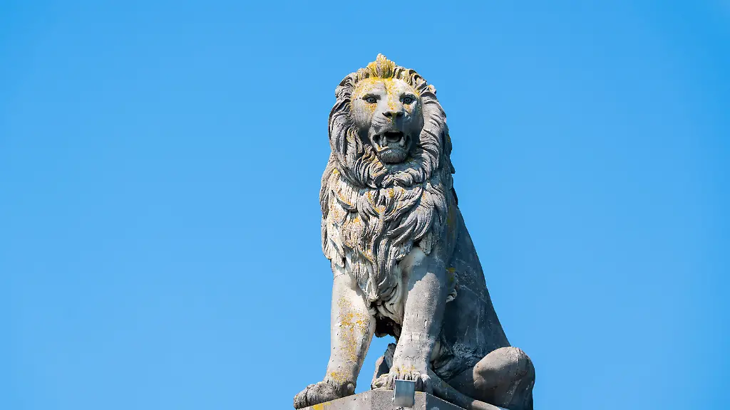 Der-Bayerische-Loewe-an-der-Hafeneinfahrt-von-Lindau-am-Bodensee-steinerne-Statue-auf-hohem-Sockel-vor-blauem-Himmel