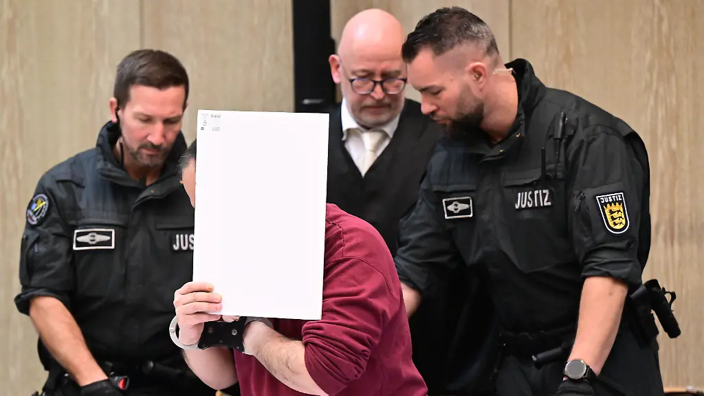 Der-Angeklagte-in-einem-Mordprozess-nach-einer-Todesfahrt-von-Mannheim-sitzt-mit-einem-Aktendeckel-vor-seinem-Gesicht-im-Gerichtssaal-rechts-neben-ihm-steht-sein-Verteidiger-Uwe-Kosmala-Bei-der-Fahrt-Anfang-Maerz-war-ein-Mann-laut-Staatsanwaltschaft-gezielt-auf-Menschen-zugefahren-Eine-83-jaehrige-Frau-und-ein-54-jaehriger-Mann-wurden-getoetet-14-Menschen-wurden-verletzt-fuenf-von-ihnen-schwer