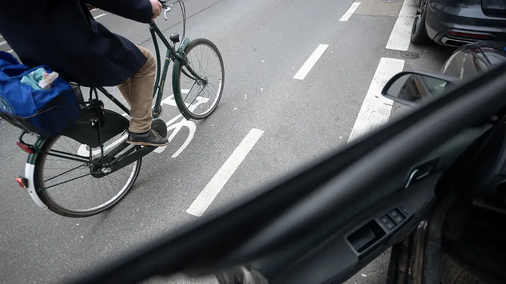 Ein-Fahrradfahrer-faehrt-auf-einer-Fahrradstrasse-an-einem-Schutzstreifen-fuer-geoeffnete-Tueren-an-Autos-vorbei-Der-Schutzstreifen-soll-verhindern-dass-Fahrradfahrer-durch-unachtsam-geoeffnete-Tueren-verletzt-werden
