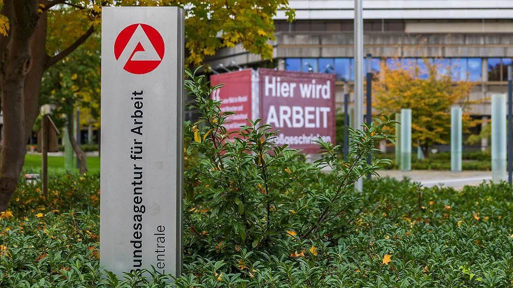 Aussenaufnahme-des-Eingangsbereichs-der-Zentrale-der-Bundesagentur-fuer-Arbeit-in-Nuernberg-Bayern-Deutschland-Im-Vordergrund-ist-ein-Schild-mit-dem-Logo-und-Schriftzug-der-Behoerde-zu-sehen-Im-Hintergrund-befindet-sich-ein-Plakat-mit-der-Aufschrift-Hier-wird-ARBEIT-beschrieben-Die-Umgebung-ist-herbstlich-mit-gefaerbtem-Laub-und-Straeuchern-Bundesagentur-fuer-Arbeit-im-Herbst-Nuernberg-17-10