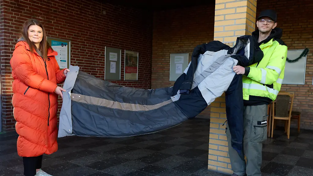 PRODUKTION-15-12-2025-Schleswig-Holstein-Elmshorn-Anna-Federmann-Leiterin-der-Bahnhofsmission-Elmshorn-und-der-Obdachlose-Pierre-Zielinski-stehen-mit-einem-sogenannten-Sheltersuit-einem-Winter-Parka-mit-integriertem-Schlafsack-im-Bahnhof-Die-Johanniter-Hilfsgemeinschaften-und-die-Sheltersuit-Foundation-starten-ihre-bundesweite-Winterhilfe-und-verteilen-ueber-800-wetterfeste-Sheltersuits-in-30-Staedten