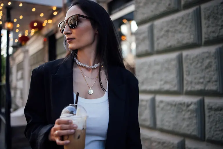 Fashionable-woman-with-dark-hair-wearing-sunglasses-and-a-black-blazer-enjoys-iced-coffee-while-strolling-in-a-vibrant-urban-environment-reflecting-contemporary-style-Modellfreigabe-vorhanden
