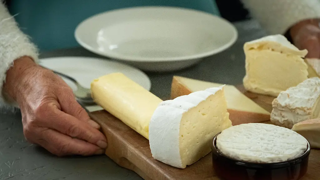 Senior-s-Hand-Serving-Gourmet-Cheese-Selection-In-this-close-up-image-the-aged-hand-of-a-senior-is-seen-delicately-serving-a-selection-of-fine-cheeses-The-assortment-which-includes-various-textures-and-rinds-is-presented-on-a-wooden-cutting-board-beside-clean-white-plates-hinting-at-an-elegant-dining-experience-The-cozy-and-inviting-atmosphere-is-underscored-by-the-soft-focused-lighting-on-the-hand-and-cheese-Senior-s-Hand-Serving-Gourmet-Cheese-Selection-High-quality-photo-Copyright-xZoonar
