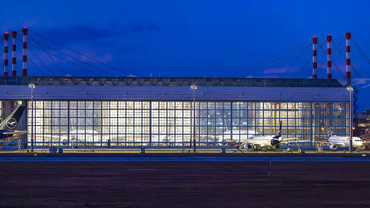 Ein-Hangar-am-Flughafen-Muenchen-ist-in-den-Abendstunden-hell-erleuchtet-Lufthansa-Flugzeuge-stehen-vor-und-in-der-Wartungshalle-waehrend-die-ikonischen-rot-weiss-gestreiften-Schornsteine-der-Flughafeninfrastruktur-im-Hintergrund-sichtbar-sind-Die-Szenerie-wird-durch-die-tiefblaue-Daemmerung-und-die-kuenstliche-Beleuchtung-gepraegt-verdi-Warnstreik-am-Flughafen-Muenchen-27-02