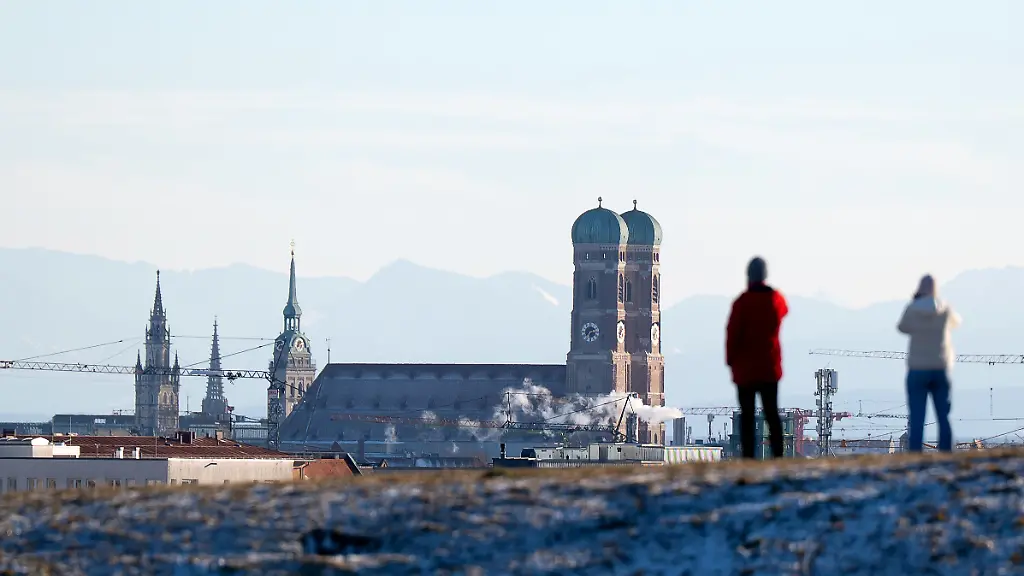 Ein-Mann-und-eine-Frau-nutzen-das-schoene-Wetter-im-Olympiapark-fuer-einen-Spaziergang-Im-Hintergrund-sind-die-Tuerme-des-Rathauses-l-r-der-Kirche-Alter-Peter-und-der-Frauenkirche-zu-sehen