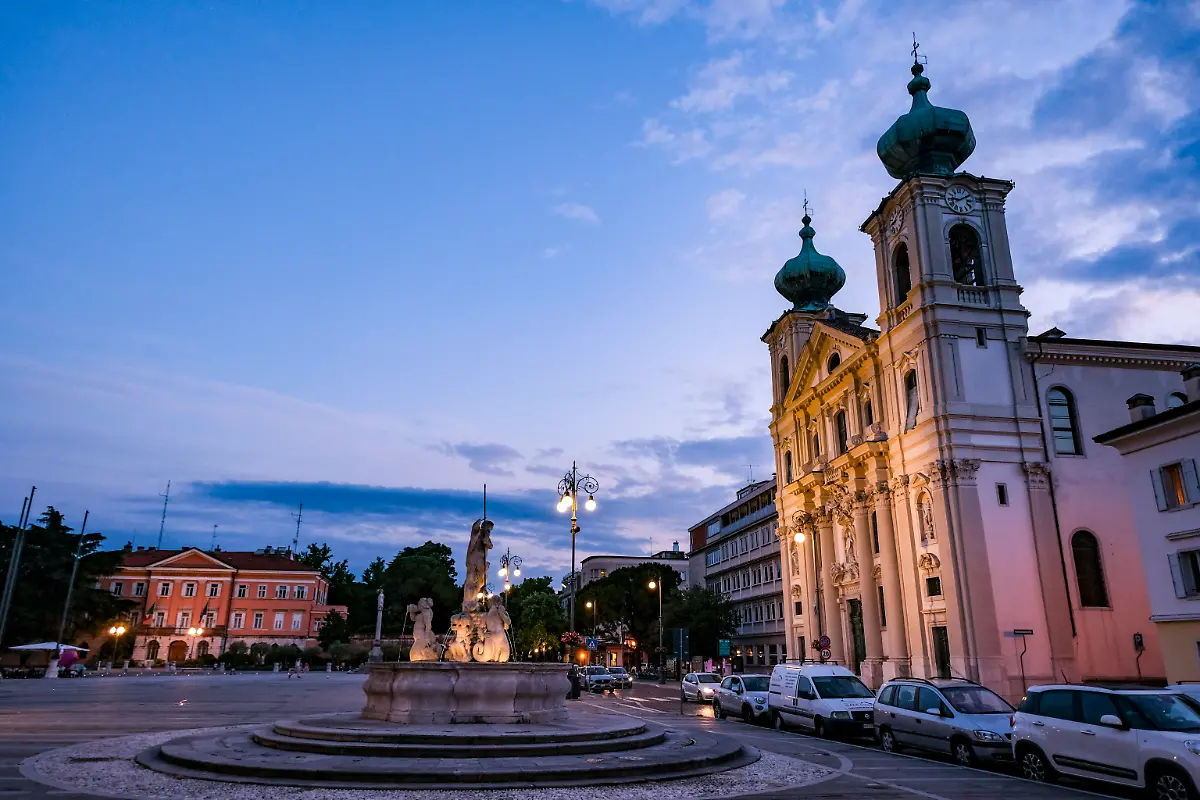 Die-Piazza-della-Vittoria-mit-der-Jesuiten-Kirche-Sant-Ignazio-und-dem-Neptun-Brunnen-am-Abend-in-der-Daemmerung-Das-Staedtepaar-Gorizia-Italien-und-Nova-Gorica-Slowenien-begeht-das-Jahr-2025-als-Europaeische-Kulturhauptstadt-gemeinsam-Erstmals-praesentiert-sich-ein-grenzuebergreifendes-Staedtepaar-als-europaeische-Kulturmetropole-Das-Motto-der-Europaeischen-Kulturhauptstadt-2025-heisst-Go-Borderless
