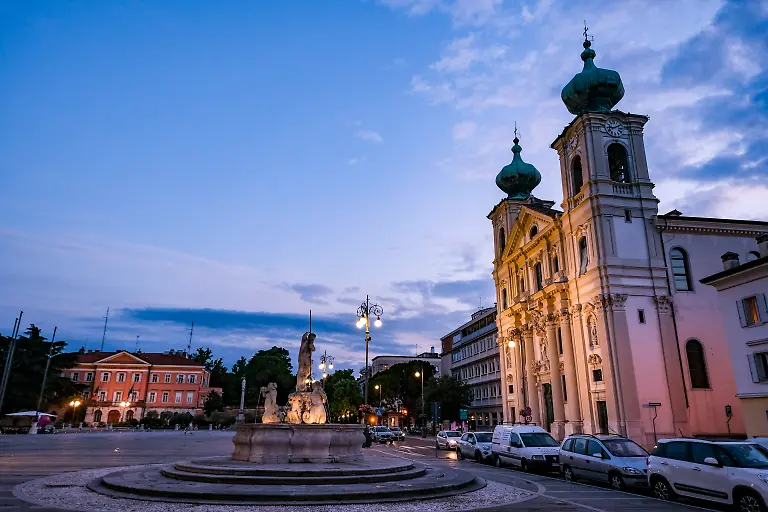 Die-Piazza-della-Vittoria-mit-der-Jesuiten-Kirche-Sant-Ignazio-und-dem-Neptun-Brunnen-am-Abend-in-der-Daemmerung-Das-Staedtepaar-Gorizia-Italien-und-Nova-Gorica-Slowenien-begeht-das-Jahr-2025-als-Europaeische-Kulturhauptstadt-gemeinsam-Erstmals-praesentiert-sich-ein-grenzuebergreifendes-Staedtepaar-als-europaeische-Kulturmetropole-Das-Motto-der-Europaeischen-Kulturhauptstadt-2025-heisst-Go-Borderless