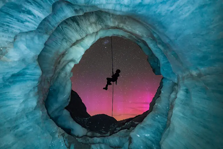I-originally-found-this-ice-cave-called-a-moulin-8-months-prior-to-setting-up-this-shot-in-Aoraki-Mount-Cook-National-Park-Glaciers-are-a-very-dynamic-environment-so-I-kept-going-back-to-monitor-the-changes-of-this-moulin-over-the-8-month-period-As-the-opening-of-the-cave-formed-I-envisioned-setting-up-a-night-shot-with-my-friend-abseiling-down-the-mouth-of-the-cave-with-New-Zealand-s-amazing-starry-sky-in-the-background-One-magical-night-everything-finally-came-together-To-my-surprise-the-Aurora-Australis-also-lit-up-the-sky-I-managed-to-capture-my-friend-s-silhouette-perfectly-placed-in-the-center-of-the-cave-s-opening-and-I-love-how-the-pinky-tones-of-the-aurora-contrast-with-the-icy-colors-of-the-cave
