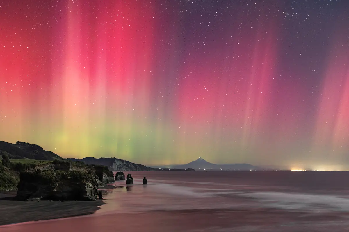 A-rare-aurora-event-lit-the-west-coast-of-New-Zealand-s-North-Island-in-vivid-curtains-of-pink-and-green-In-the-foreground-the-rock-formations-known-as-the-Three-Sisters-stand-as-guardians-of-the-shoreline-while-the-sacred-Taranaki-Maunga-rises-in-the-distance-Within-Te-ao-Maori-the-Maori-worldview-such-natural-features-are-not-just-landscapes-but-ancestral-presences-carrying-the-role-of-guardianship-Beneath-the-aurora-earth-sky-and-ancestry-converge-in-a-moment-both-fleeting-and-timeless-After-capturing-my-first-aurora-during-the-May-2024-storm-I-was-hooked-Travelling-several-hours-from-my-home-to-this-special-location-I-hoped-the-forecasts-would-be-correct-Even-with-a-near-full-moon-the-powerful-display-shone-across-the-sky-I-could-see-the-beams-dancing-overhead-a-truly-spectacular-sight