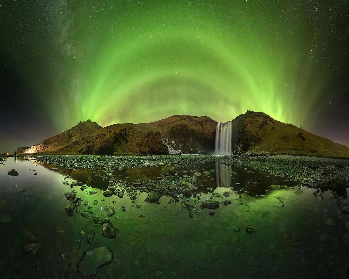 On-nights-like-this-Iceland-feels-otherworldly-The-aurora-unfolded-in-perfectly-layered-arcs-painting-the-entire-valley-with-shades-of-emerald-while-the-waterfall-echoed-under-the-glow-The-reflection-on-the-frozen-shallows-completed-the-symmetry-making-the-whole-landscape-look-like-a-portal-to-another-world-It-s-impossible-to-stand-here-and-not-feel-the-power-of-nature-When-you-re-standing-before-this-waterfall-your-first-impulse-is-to-focus-on-it-using-focal-lengths-around-20mm-However-when-I-observed-the-scene-I-noticed-that-the-Aurora-Borealis-was-forming-arcs-aligned-with-the-mountains-surrounding-the-waterfall-so-I-opted-to-take-a-panoramic-image-using-a-12mm-fisheye-lens-to-reduce-the-number-of-images-needed-to-cover-the-entire-scene-I-also-chose-to-take-advantage-of-the-stream-formed-by-the-waterfall-to-capture-the-reflection-of-the-landscape-and-the-sky-thus-adding-more-complexity-to-the-composition-Wearing-waterproof-boots-or-at-least-what-should-have-been-I-entered-the-watercourse-set-up-my-tripod-very-low-and-took-the-sequence-of-images-to-assemble-the-panorama-later