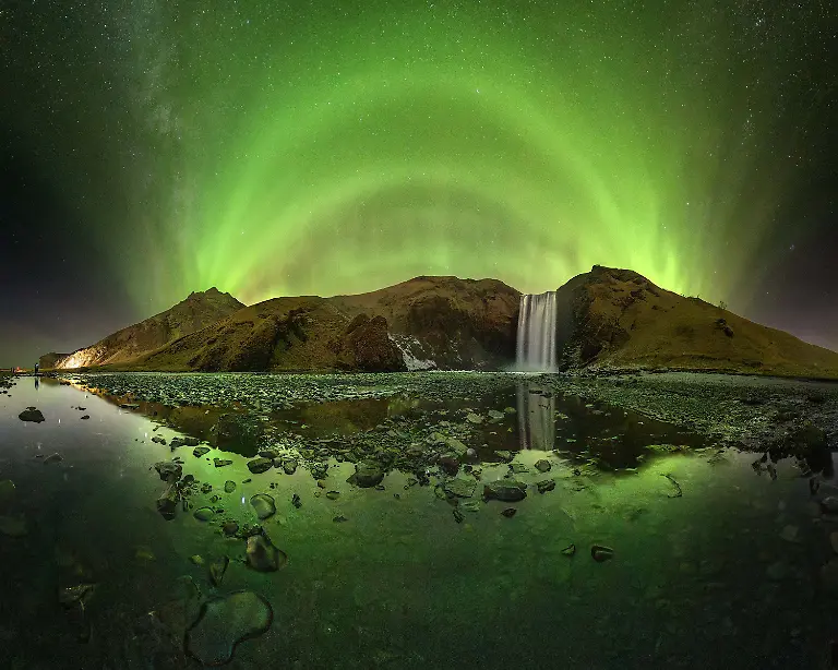 On-nights-like-this-Iceland-feels-otherworldly-The-aurora-unfolded-in-perfectly-layered-arcs-painting-the-entire-valley-with-shades-of-emerald-while-the-waterfall-echoed-under-the-glow-The-reflection-on-the-frozen-shallows-completed-the-symmetry-making-the-whole-landscape-look-like-a-portal-to-another-world-It-s-impossible-to-stand-here-and-not-feel-the-power-of-nature-When-you-re-standing-before-this-waterfall-your-first-impulse-is-to-focus-on-it-using-focal-lengths-around-20mm-However-when-I-observed-the-scene-I-noticed-that-the-Aurora-Borealis-was-forming-arcs-aligned-with-the-mountains-surrounding-the-waterfall-so-I-opted-to-take-a-panoramic-image-using-a-12mm-fisheye-lens-to-reduce-the-number-of-images-needed-to-cover-the-entire-scene-I-also-chose-to-take-advantage-of-the-stream-formed-by-the-waterfall-to-capture-the-reflection-of-the-landscape-and-the-sky-thus-adding-more-complexity-to-the-composition-Wearing-waterproof-boots-or-at-least-what-should-have-been-I-entered-the-watercourse-set-up-my-tripod-very-low-and-took-the-sequence-of-images-to-assemble-the-panorama-later