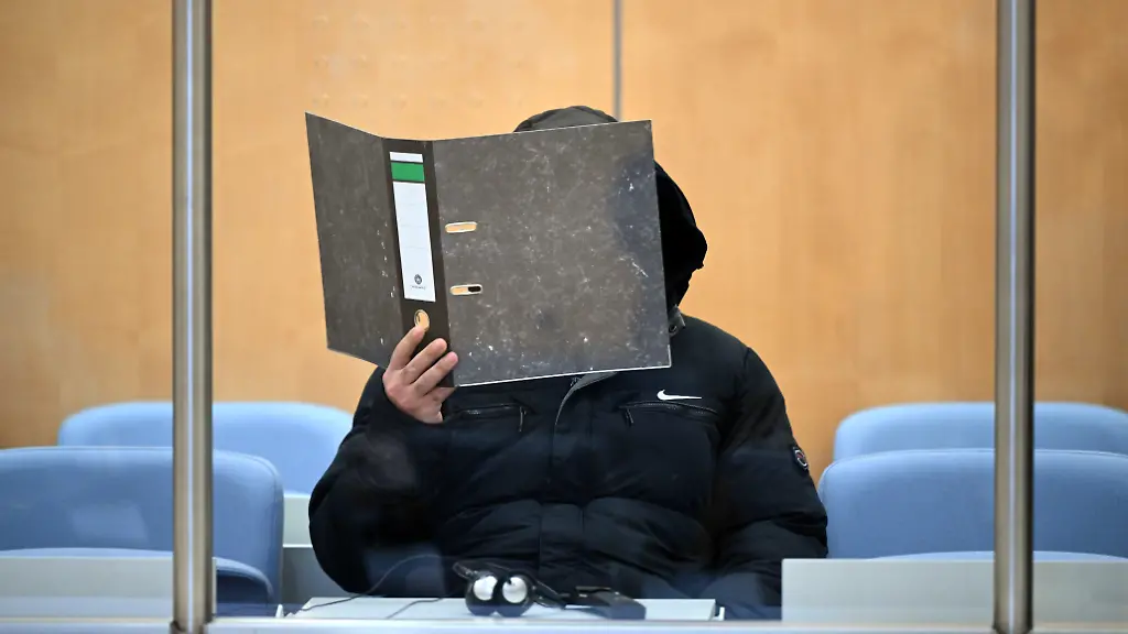 Der-Angeklagte-haelt-sich-einen-Aktenordner-vor-das-Gesicht-Ein-33-Jaehriger-aus-Monheim-muss-sich-als-mutmasslicher-frueherer-Kaempfer-der-Terrormiliz-Islamischer-Staat-am-Oberlandesgericht-verantworten-Dem-Angeklagten-werden-neben-Terrorismus-auch-Kriegsverbrechen-Verbrechen-gegen-die-Menschlichkeit-und-Beihilfe-zum-Mord-vorgeworfen